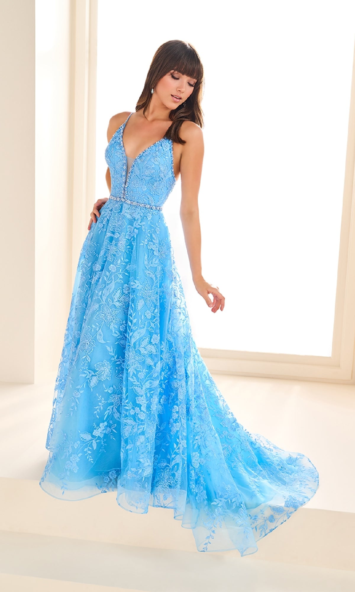 A woman stands in front of bright windows wearing the Ellie Wilde EW36088 long prom dress—a sleeveless, light blue embroidered tulle gown with a deep V-neckline, A-line skirt, and pockets.